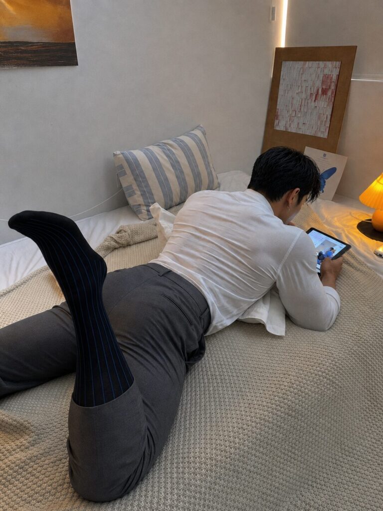 Reo lying on the bed in grey trousers and black long socks, seen from behind while looking at his phone.