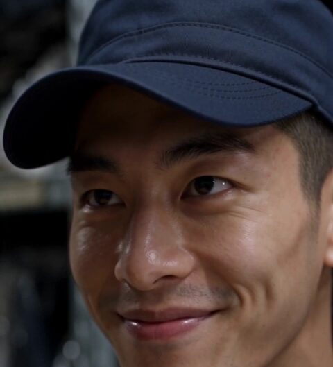 Reo looks toward the camera in his navy security cap inside the stockroom.